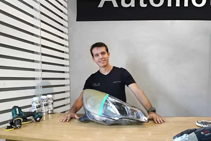 Fotografia de Amilker Kenned, proprietário da Zelo Automotivo, atrás de um farol parcialmente restaurado, mostrando a diferença entre o antes e depois.